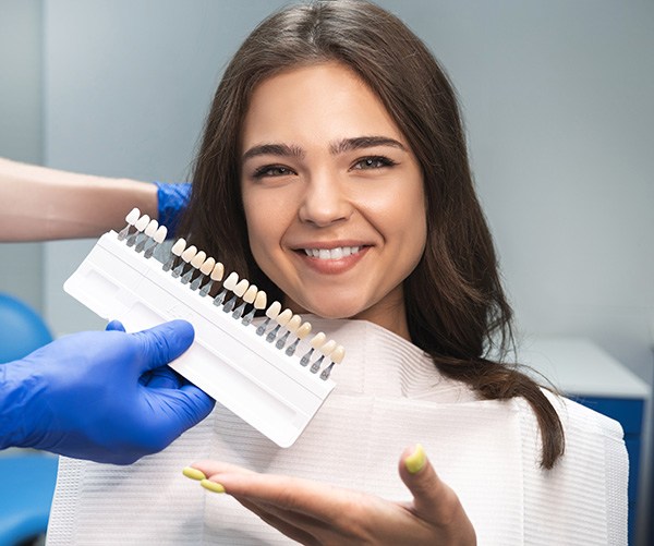 Woman with brown hair and yellow nails gesturing to shade guide held to her smile